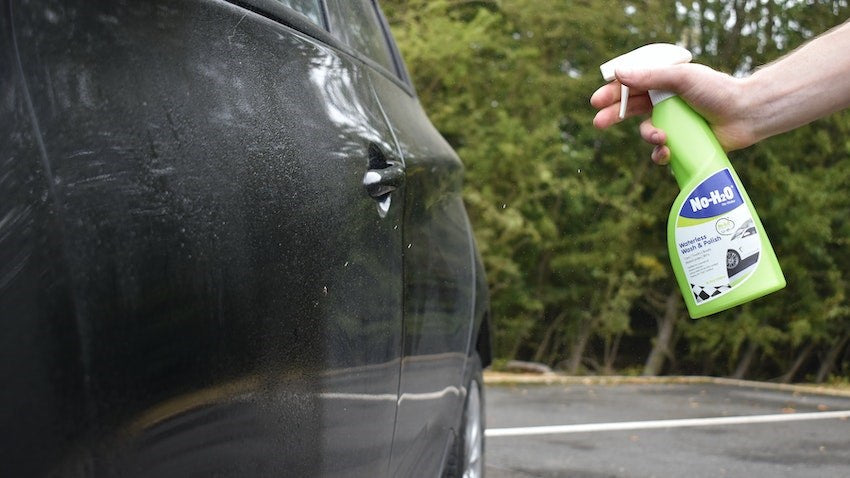 A hand sprays a green "No-H2O" bottle onto a dusty black car, demonstrating its cleaning potential in an outdoor setting.
