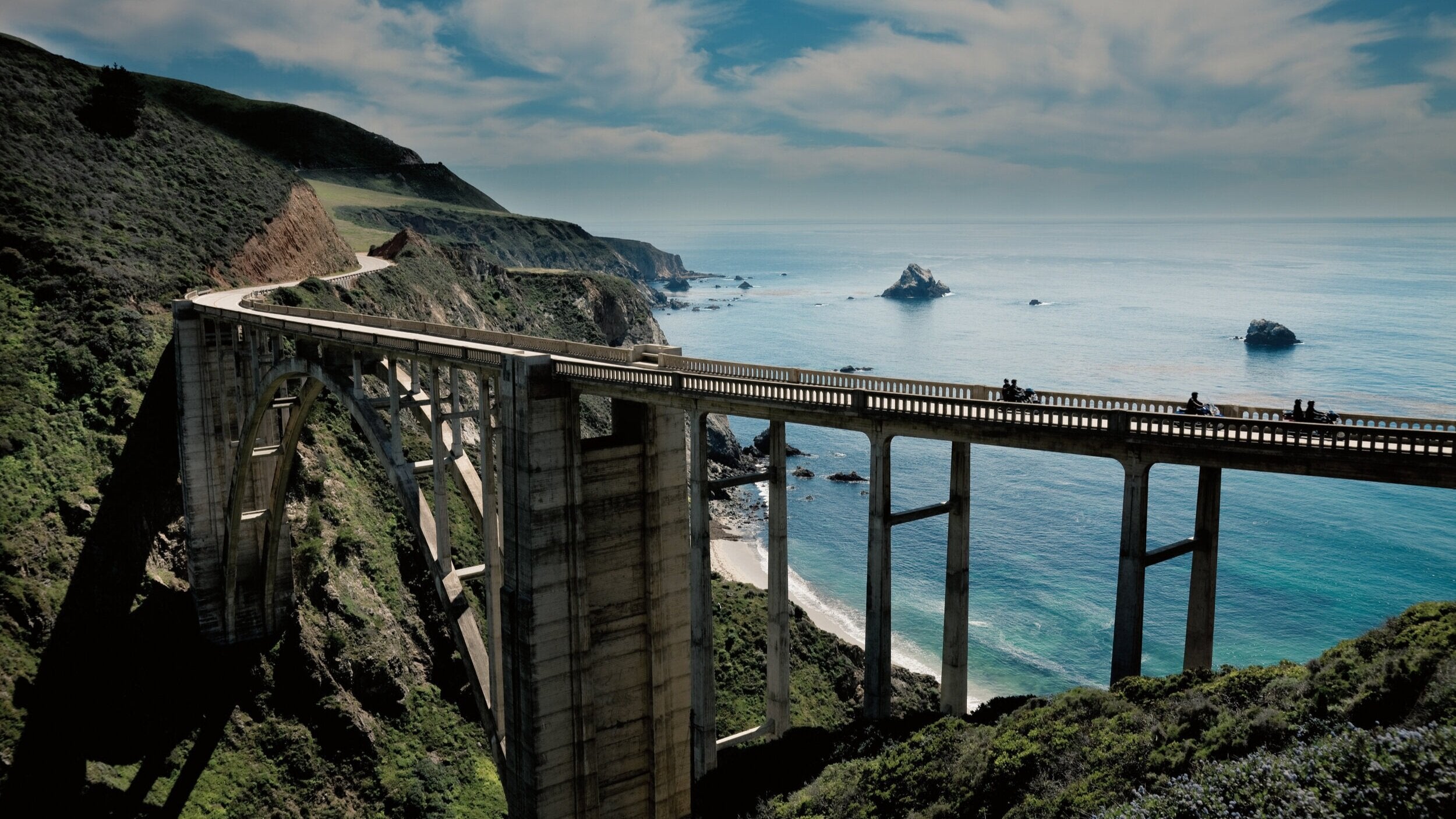 A majestic arched bridge spans a canyon, connecting verdant cliffs beside an azure sea. Silhouettes of motorcycles and riders cross the structure, contrasting the tranquil coastline and cloud-streaked sky.
